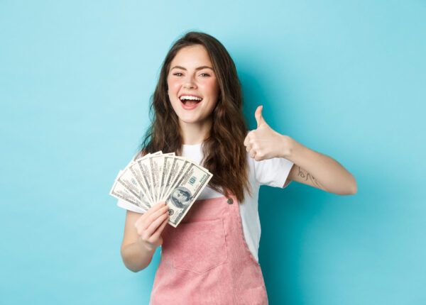 Smiling happy woman holding money, dollar bills and showing thumb up, recommending fast cash loan and looking satisfied, standing over blue background.