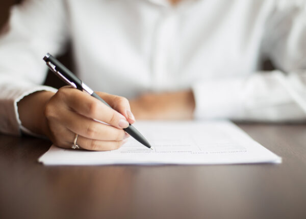Hand of young businesswoman wearing ring and sitting at table writing on document with pen in office
