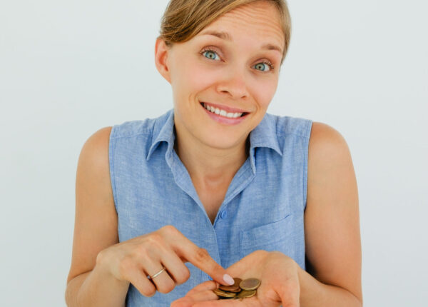 Closeup portrait of smiling young attractive woman looking at camera, shrugging and counting coins on palm. Isolated front view on grey background.
