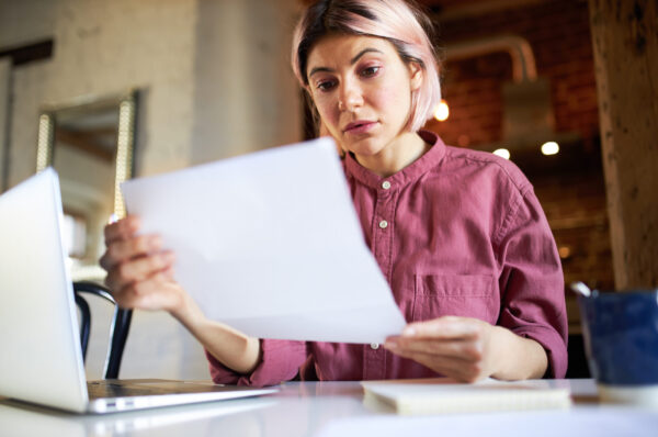 Concentrated successful young female economist analyzing important data, sitting in front of open laptop, studying accounting document. Woman office manager working from home during social distancing