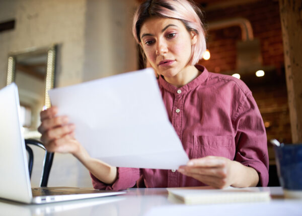 Concentrated successful young female economist analyzing important data, sitting in front of open laptop, studying accounting document. Woman office manager working from home during social distancing