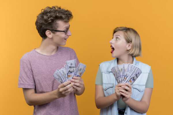 young beautiful couple wearing casual clothes man and woman showing cash looking surprised standing over orange background