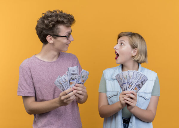 young beautiful couple wearing casual clothes man and woman showing cash looking surprised standing over orange background